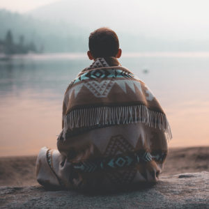A man sitting on the bank of a lake, wrapped in a blanket of Indigenous design.