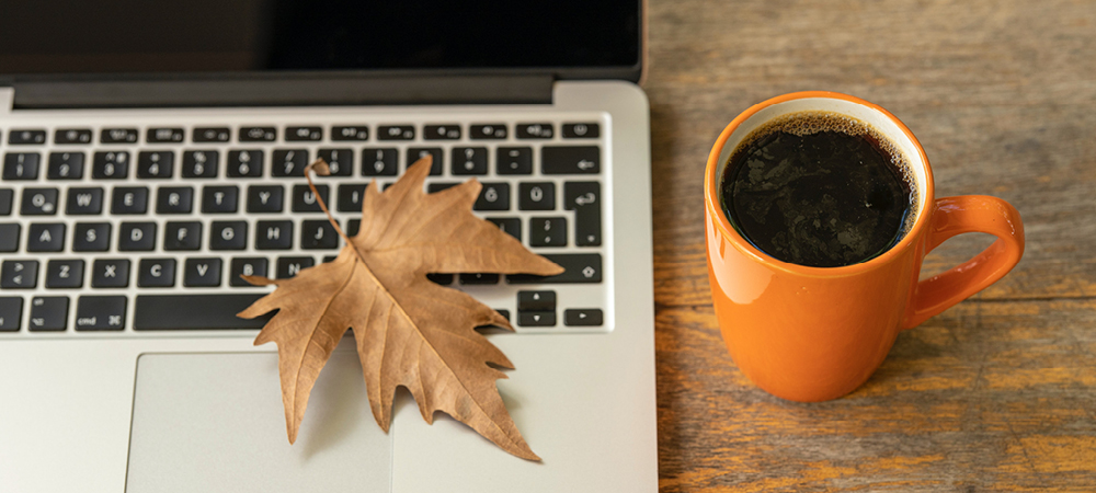 A fall leaf sitting on a laptop keyboard with an orange coffee mug alongside.