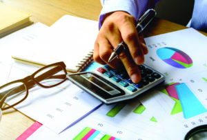 Close up of a man's hand using a calculator on a desk covered with financial data.