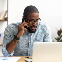 A man works at a laptop while talking on the phone.
