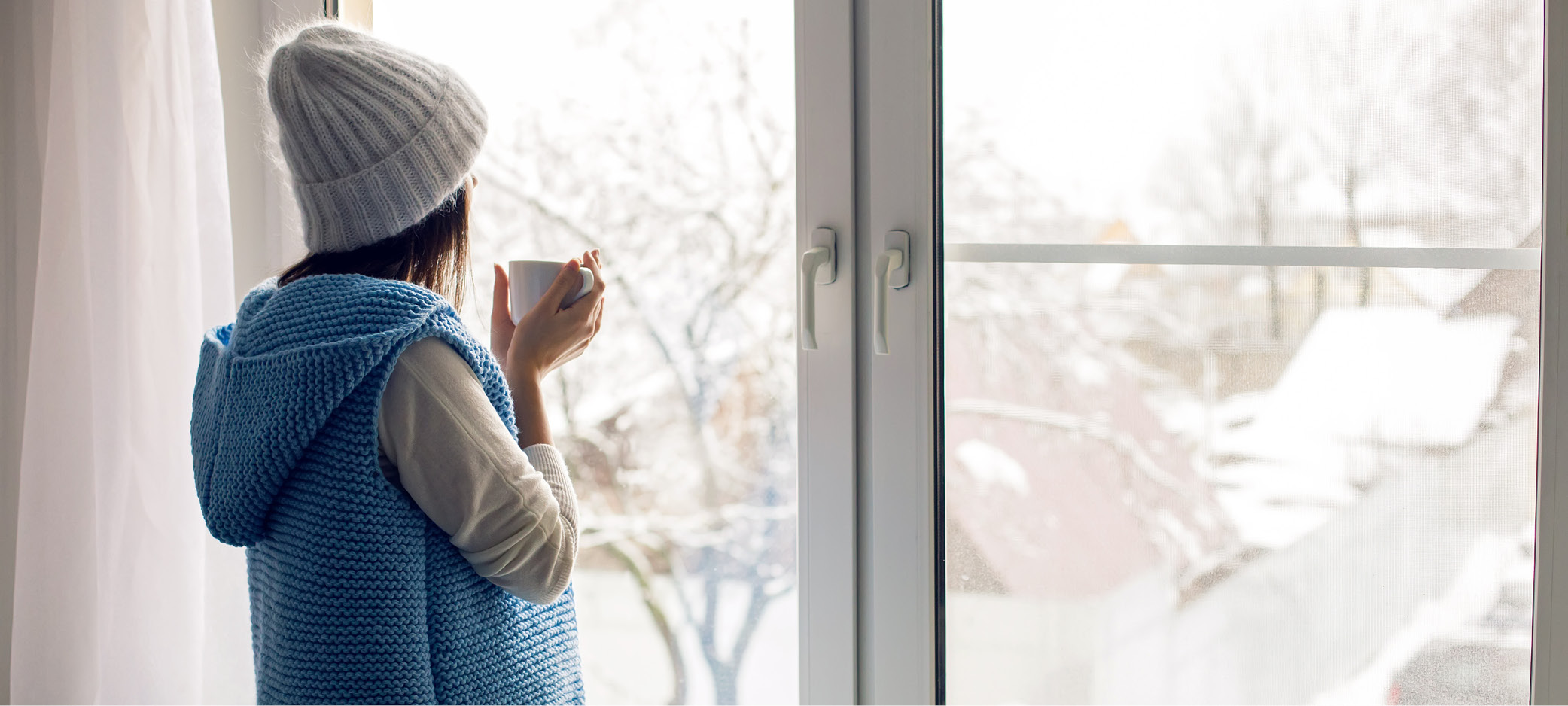 A woman holds a cup while looking out a window at a snowy residential neighbourhood