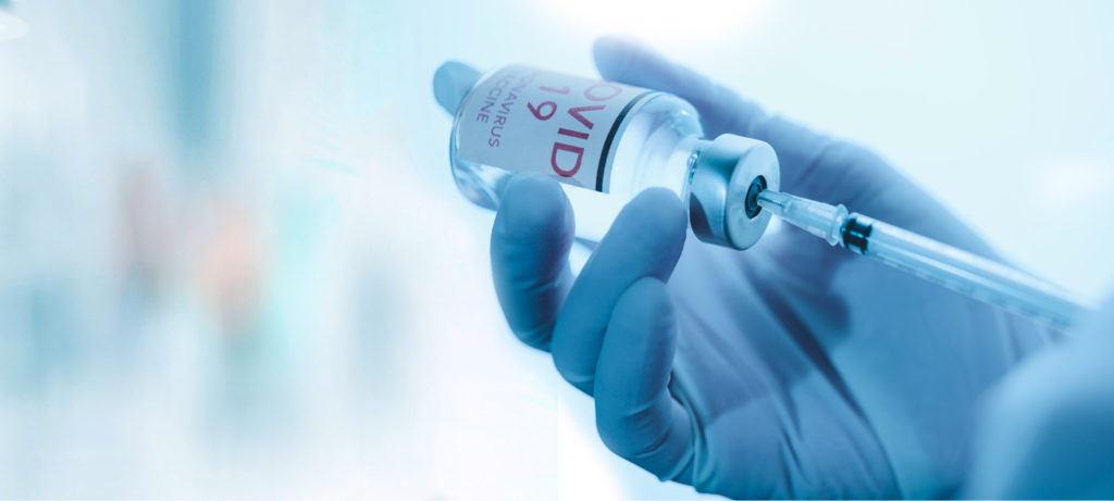 A close-up of gloved hands using a syringe to pull from a vial labeled covid-19 vaccine