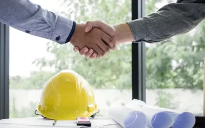 Two men shaking hands over a table with a hard hat and construction plans