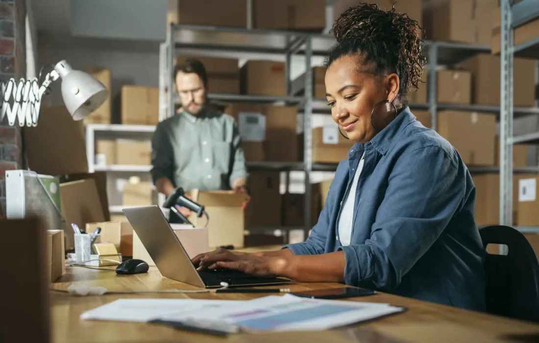 Two Warehouse Inventory Managers Talking, Using Laptop Computer and Checking Retail Stock. Rows of Shelves Full of Cardboard Box Packages in the Background.